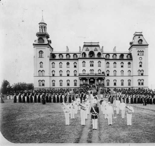 Military cadets practicing a formation outside of the Old Main. Male cadets are in the back, female cadets in the middle, and a band up front. Pictured from the east. The Old Main stood where current day Beardshear Hall stands now.