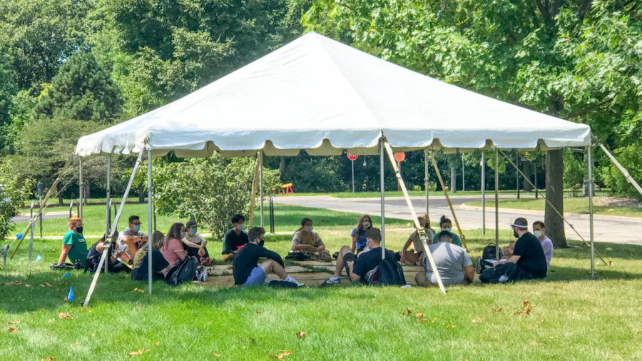 Students sit in the shade of a tent outside the design building while wearing masks and sitting 6 feet apart.