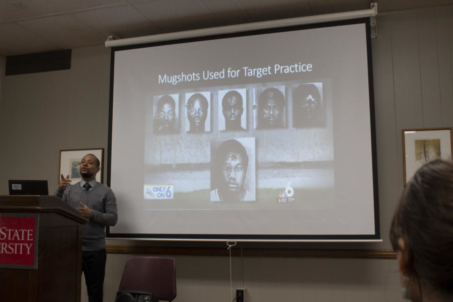 Dr. Novotny Lawrence standing in front of bullet-ridden mugshots as he discusses the past and current state of black media.