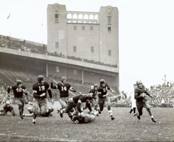 Everett Kischer carrying the ball during a football game being played before a partially filled stadium of spectators in 1938. The photo is in the digital collections of the Iowa State University Library.